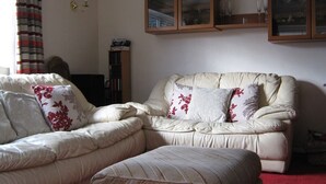 Een woonkamer met een beigekleurige zithoek, een bijpassend poefje en een roodgekleurd tapijt. Daarboven, boven de bank, staan twee houten kasten met glazen deuren.