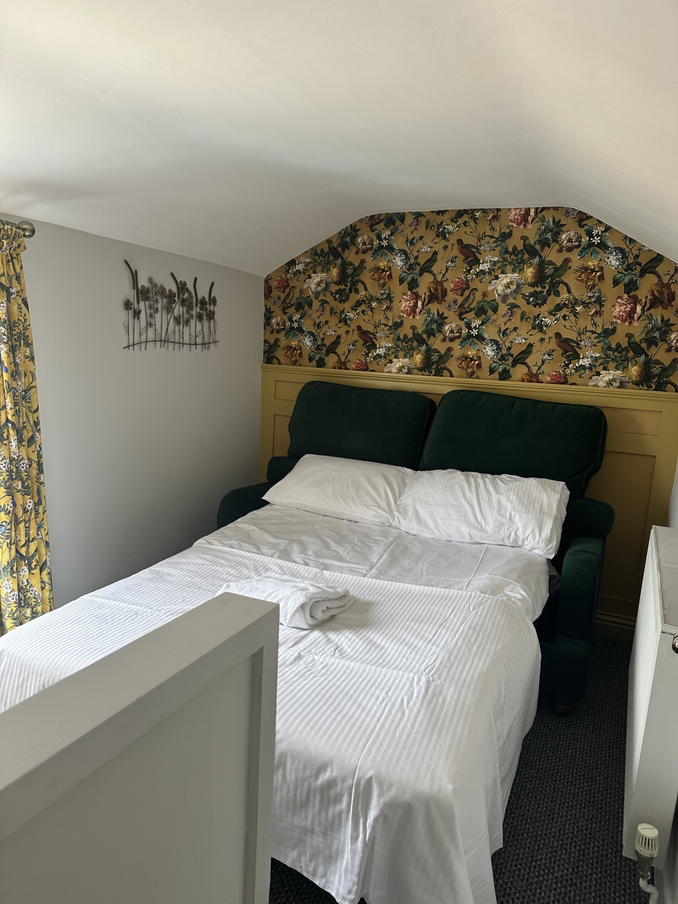 A hotel room with a bed, a headboard with floral wallpaper, a wall art piece, and a window with curtains.
