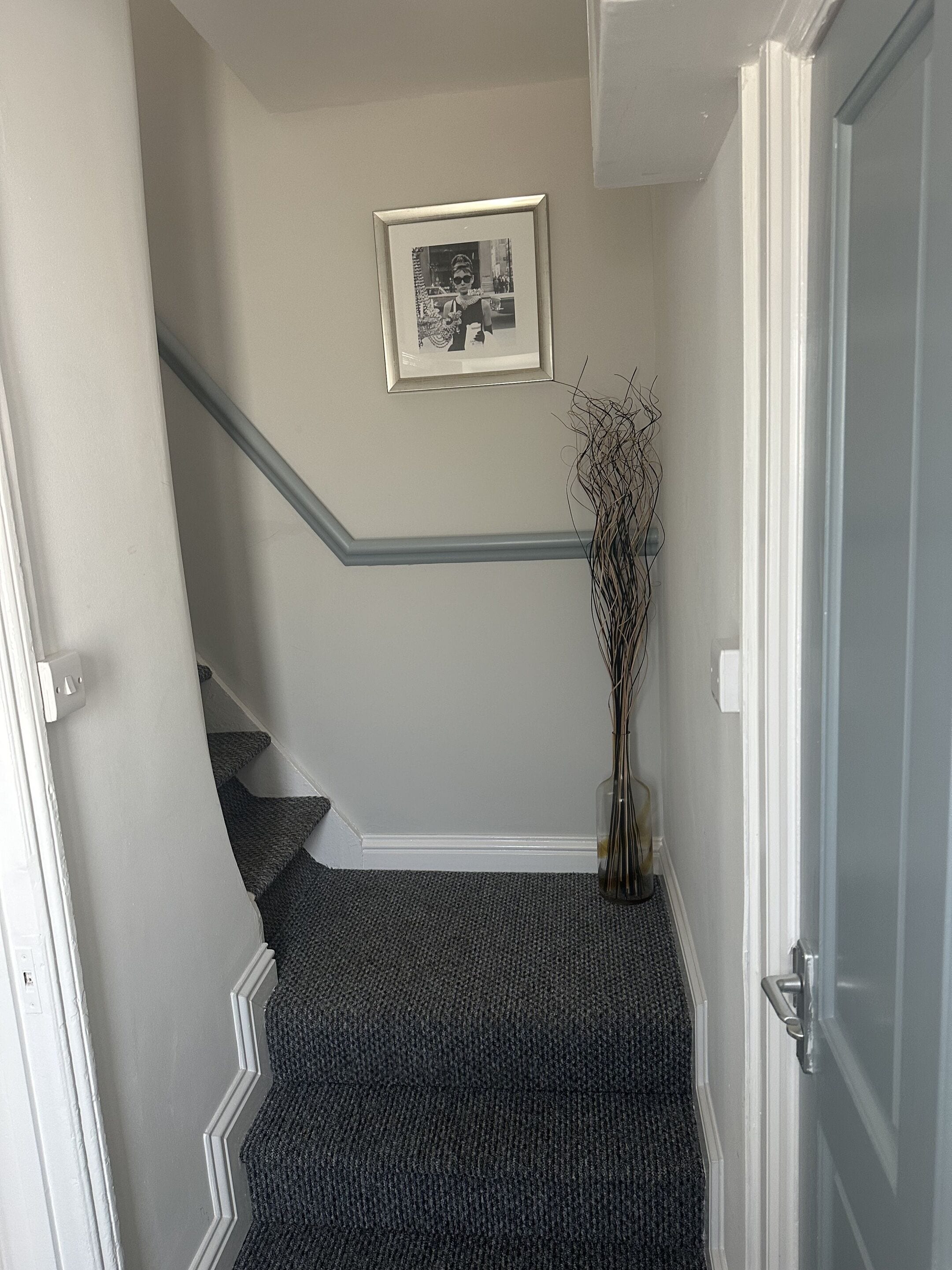 Un escalier recouvert d’une moquette à motifs, un tableau encadré accroché au mur et un vase orné de branches.