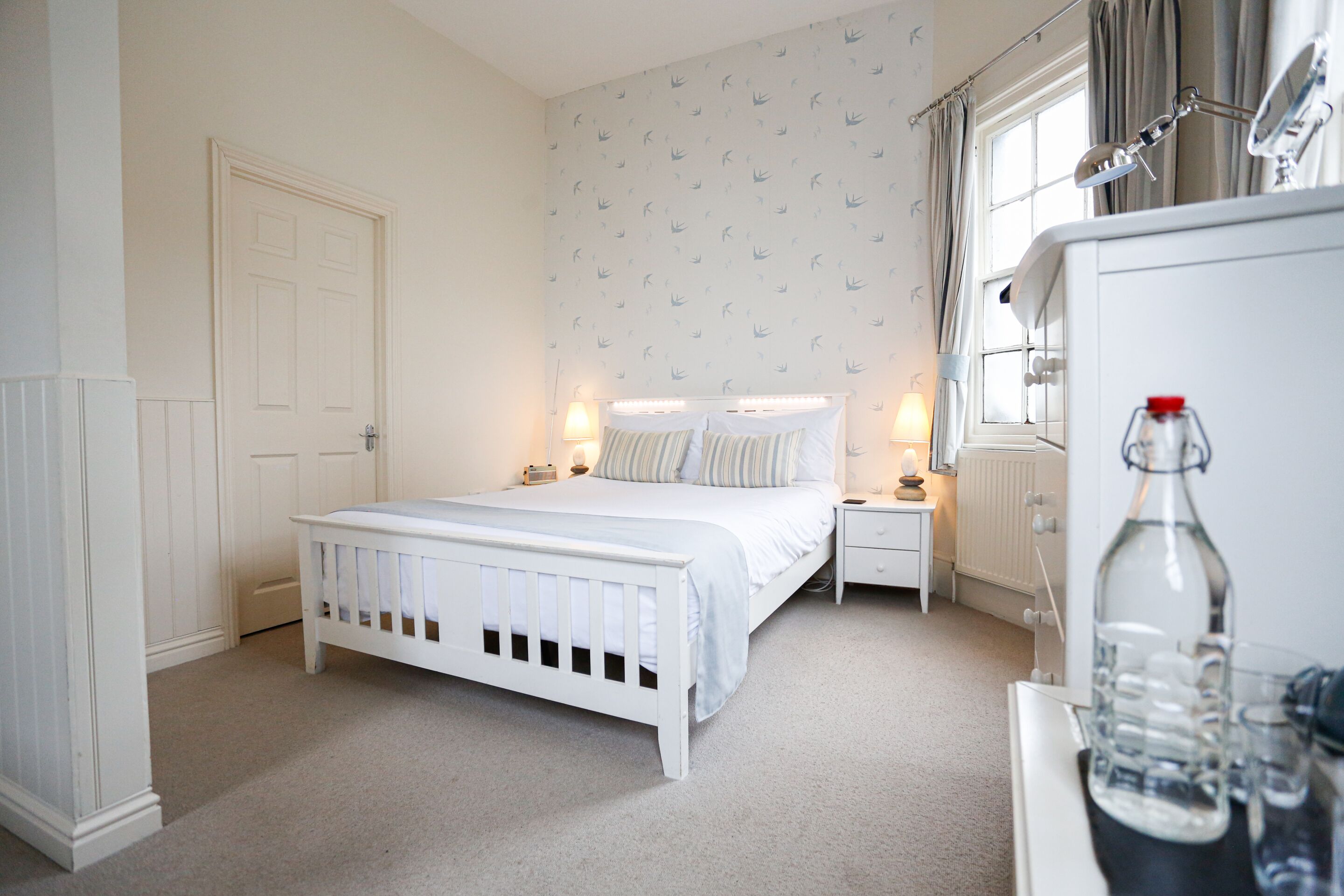 A bedroom with a white bed, bedside tables, a dresser, a mirror, and a window with curtains.