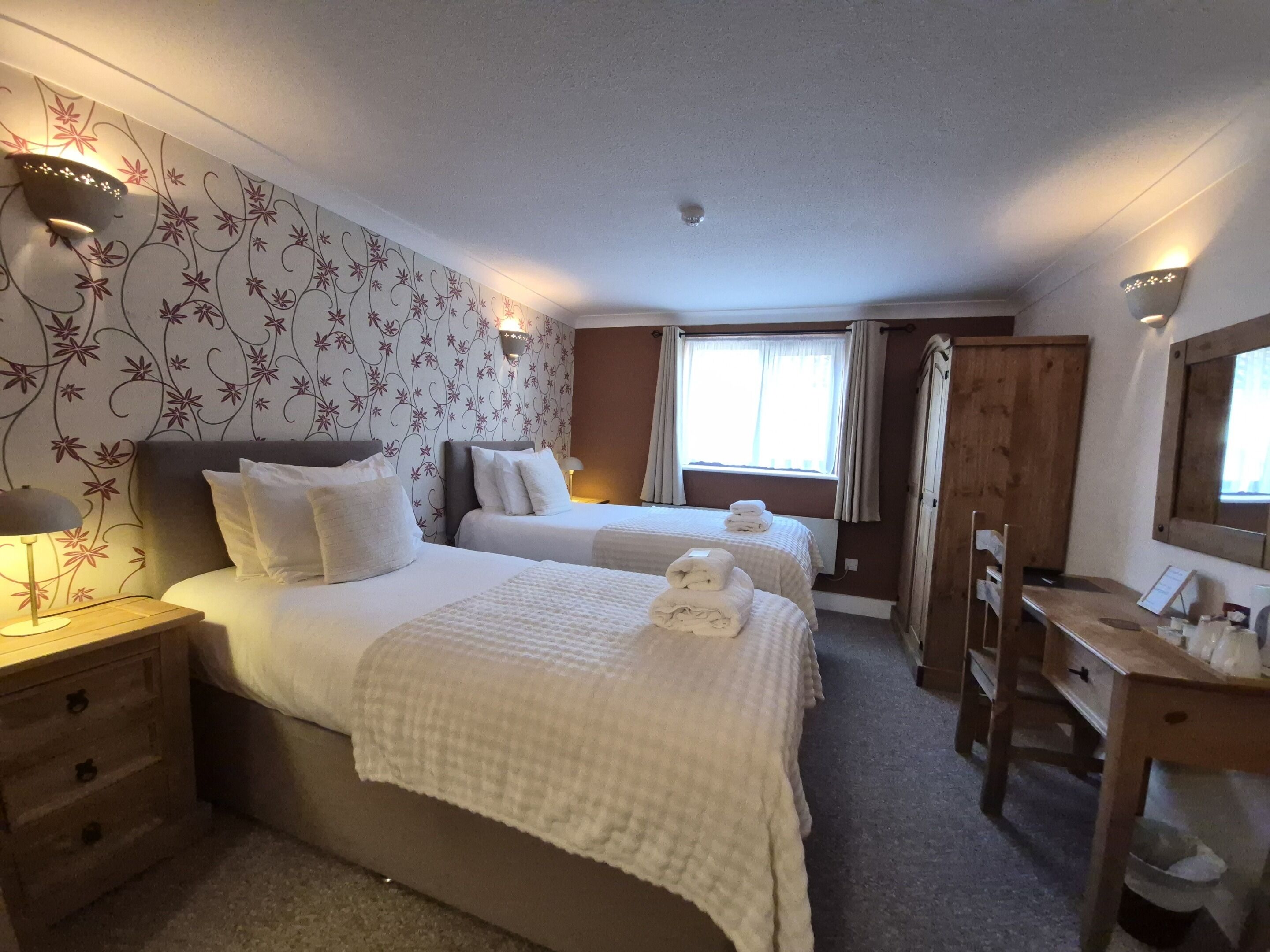 Une chambre d’hôtel avec deux lits, un bureau, une chaise, une lampe et une fenêtre avec des rideaux.