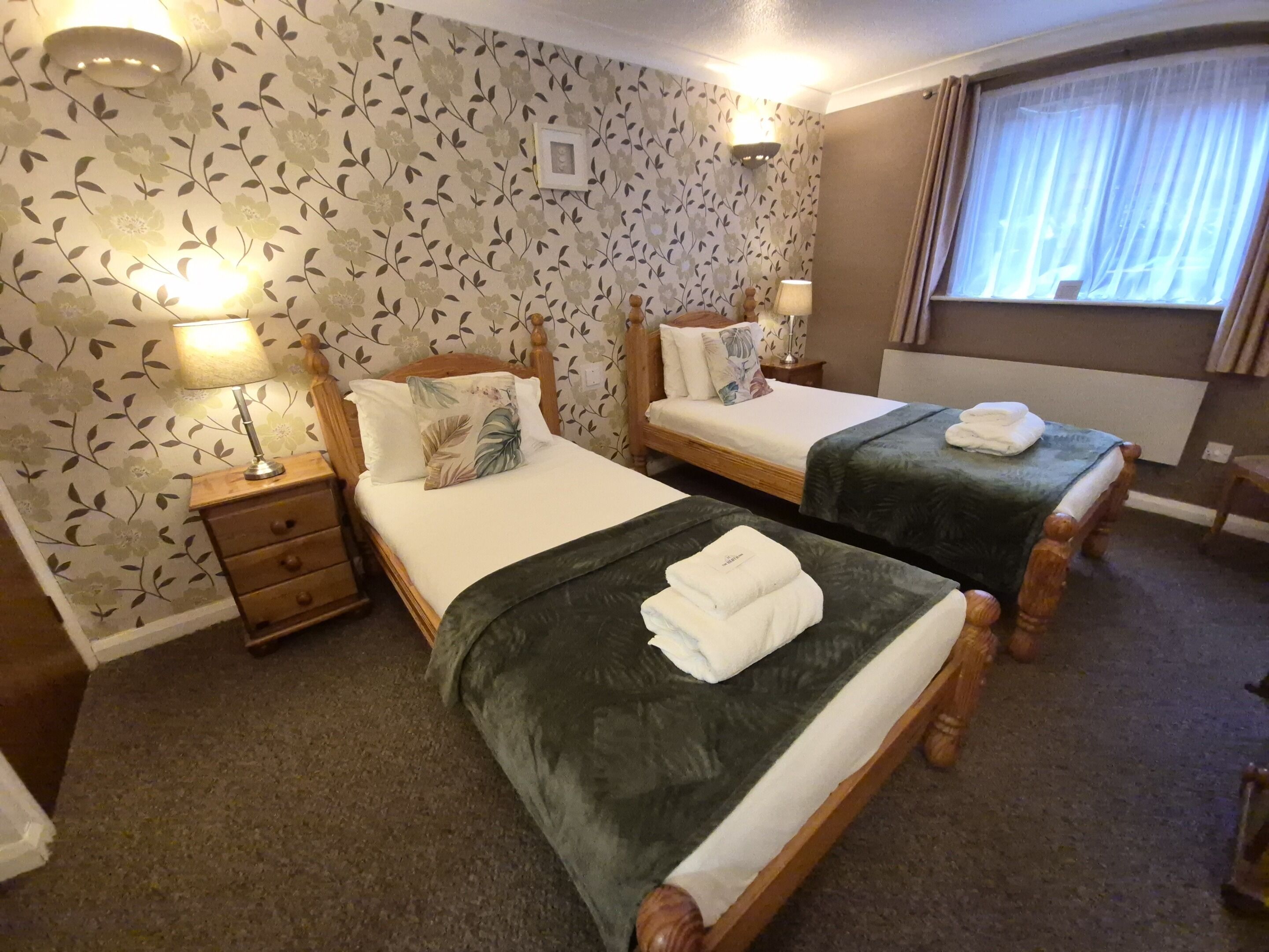 A hotel room with two beds, floral wallpaper, bedside lamps, and a window with curtains.