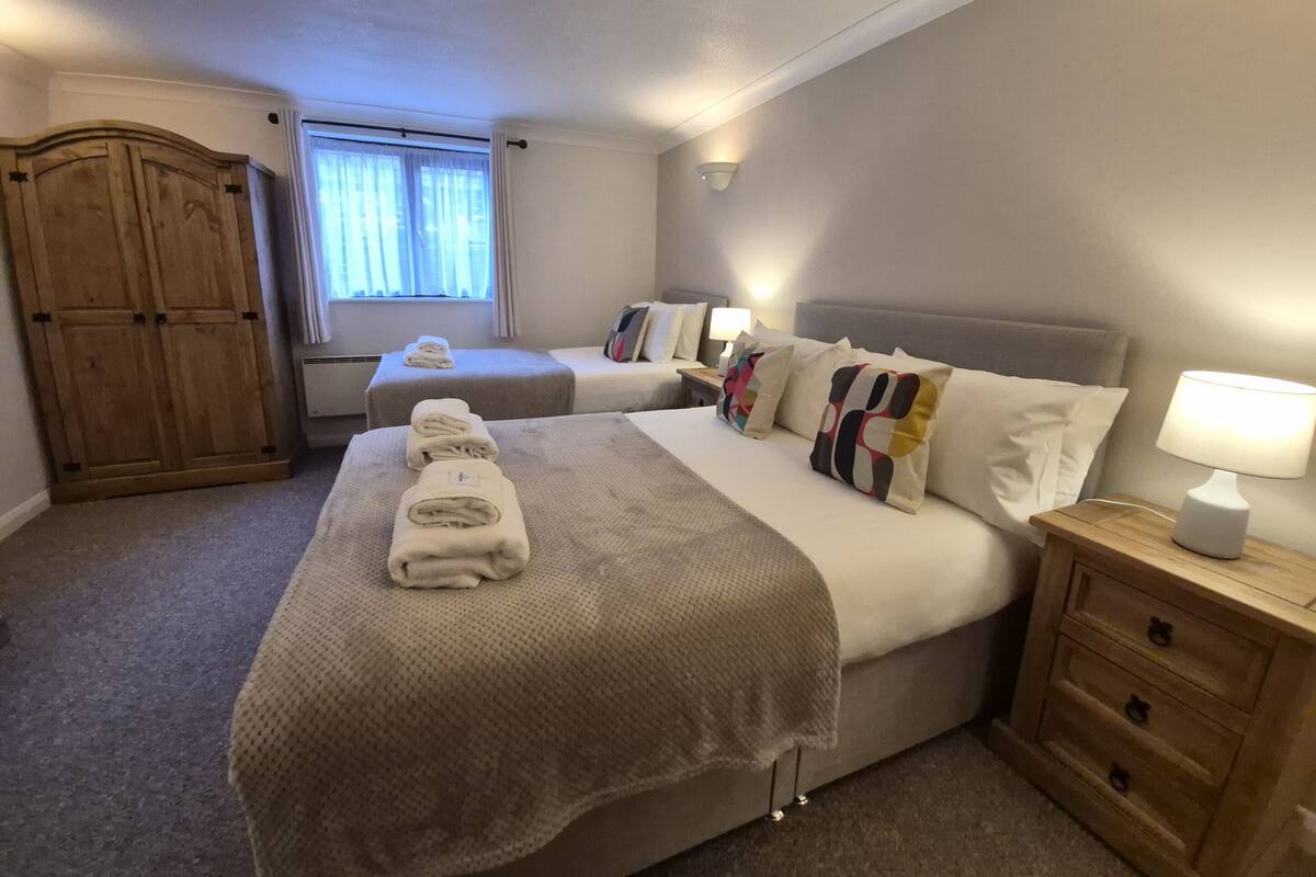 A hotel room with two beds, a wardrobe, a window with curtains, and a bedside table with a lamp.