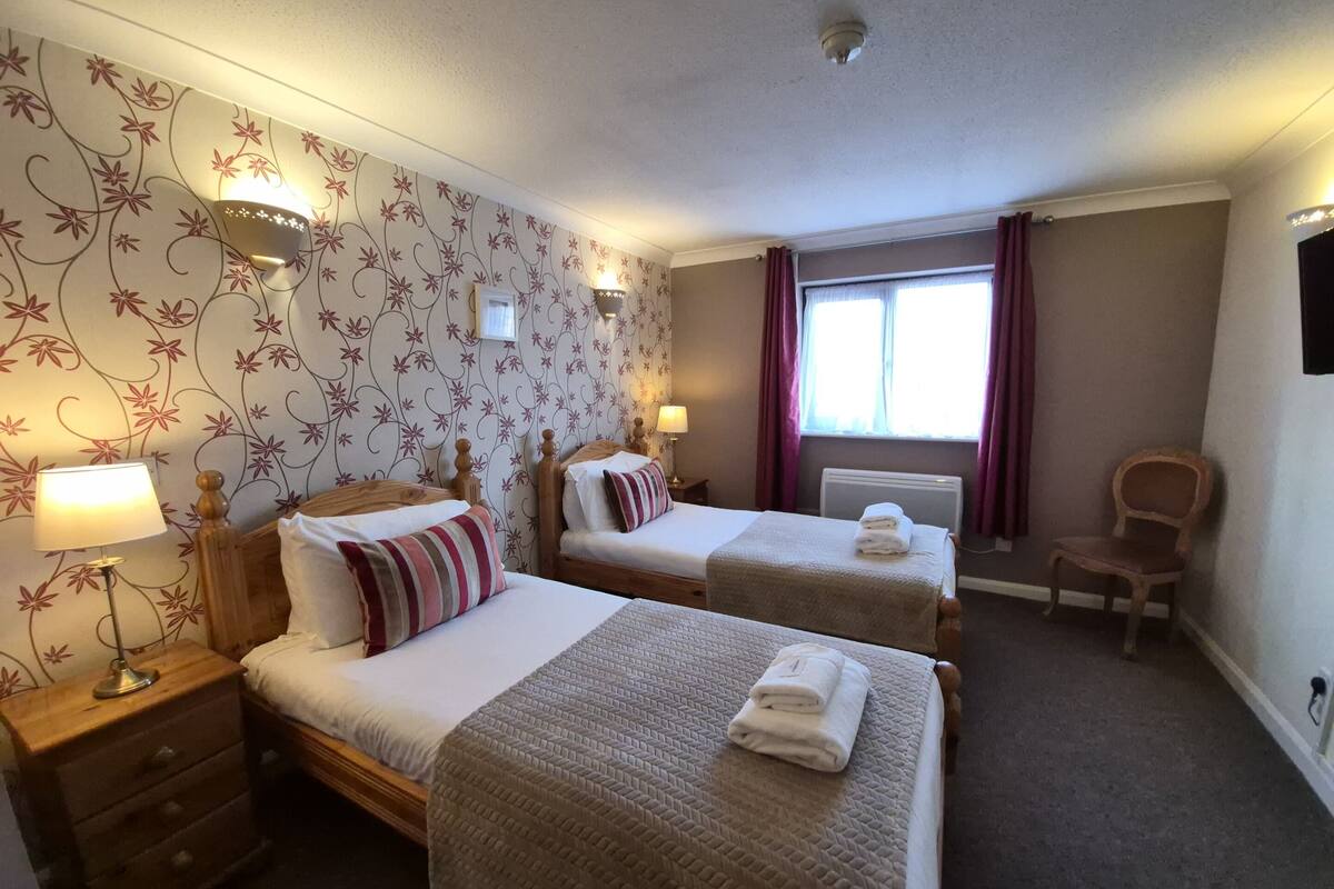A hotel room with two beds, a nightstand with a lamp, a chair, and a window with curtains.