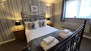 A neatly made bed with a headboard, two bedside tables with lamps, a window with curtains, and a wall with a striped pattern and a framed picture. - The Heath Inn (Leighton Buzzard)