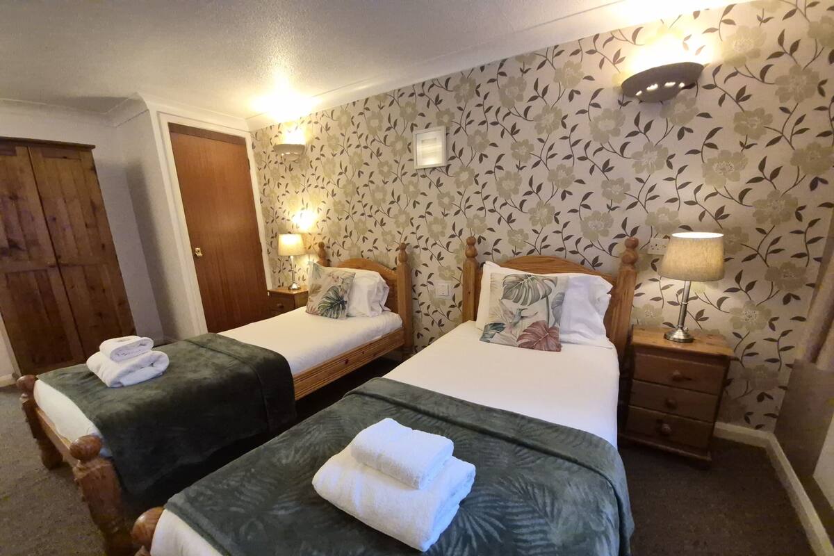 A hotel room with two beds, each with a green bedspread and white linens, a wooden headboard, and a nightstand with a lamp.