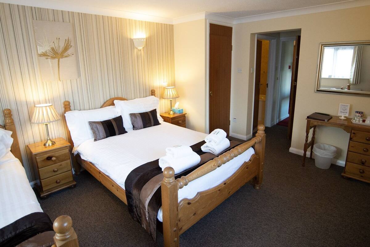 A neatly made bed with white linens and a brown headboard, a bedside lamp, a wooden nightstand, a small table with a lamp, a mirror, and a wooden dresser with drawers.