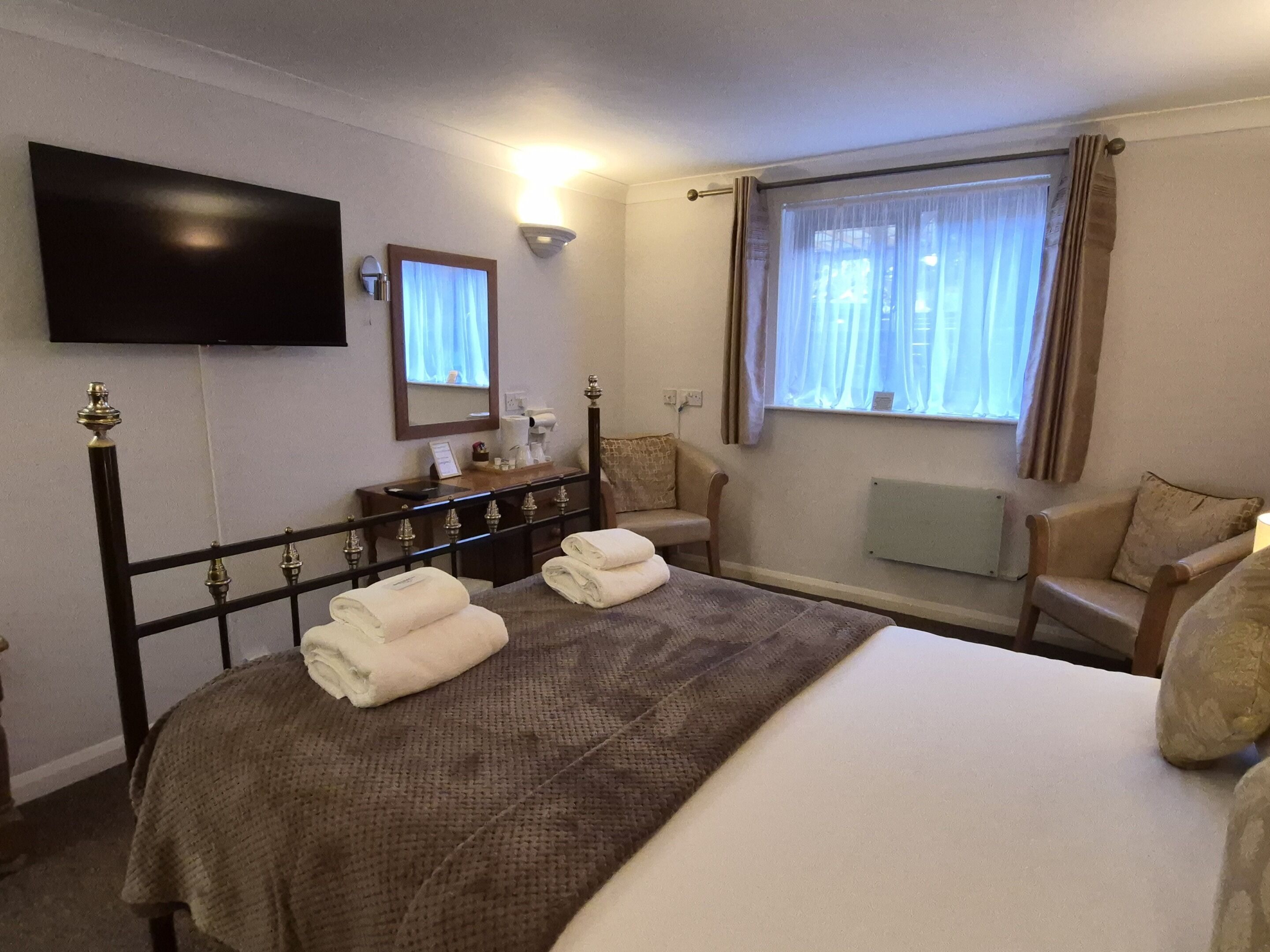 A hotel room with a bed, a TV mounted on the wall, a bedside table with a lamp, a chair, and a window with curtains.