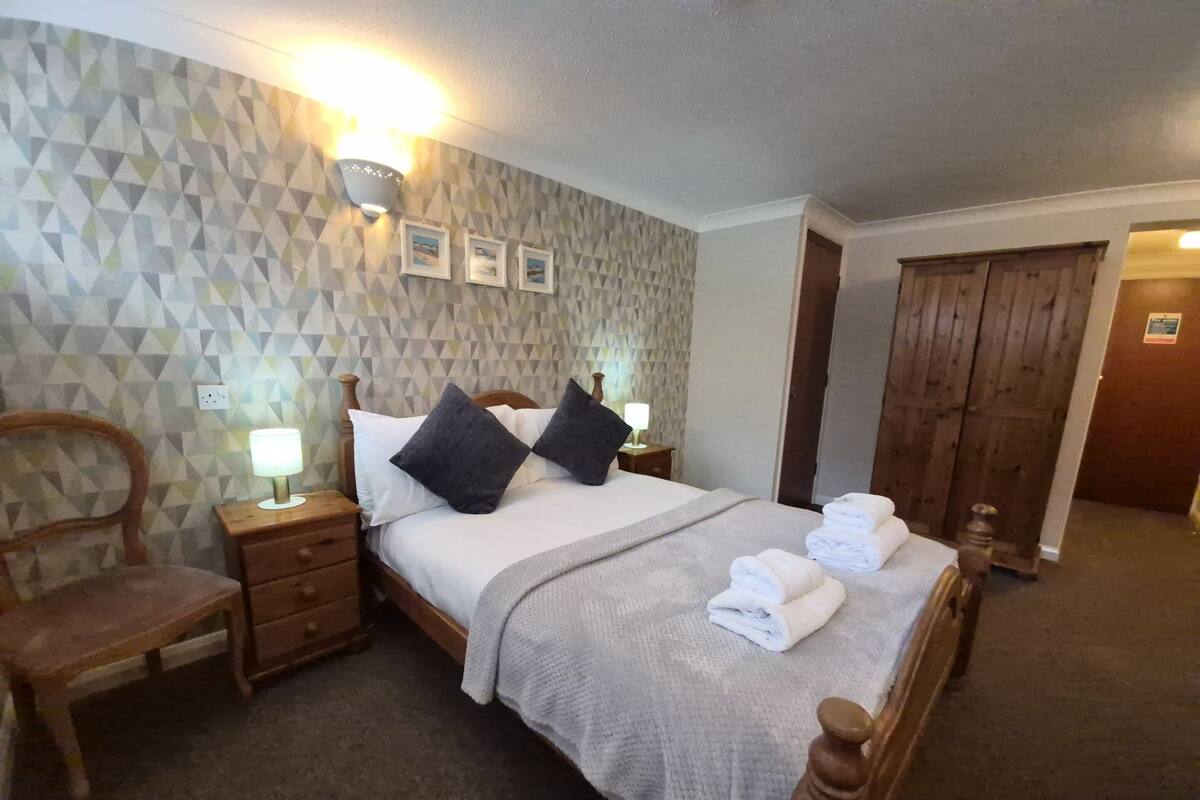 A neatly arranged hotel room with a bed, a chair, a nightstand, a lamp, and a wall with geometric patterns.