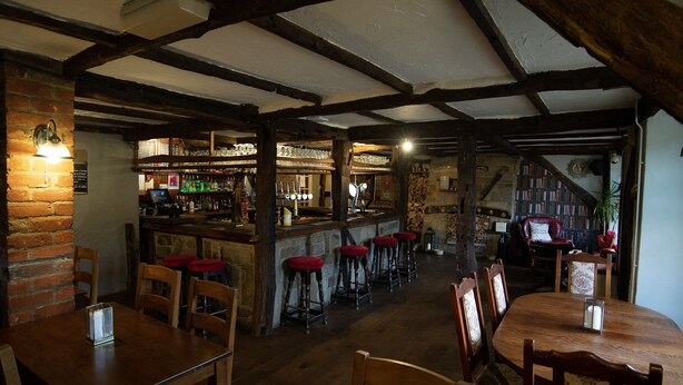 A rustic pub with exposed wooden beams, a bar area with stools, and a cozy seating arrangement. - The Heath Inn (Leighton Buzzard)