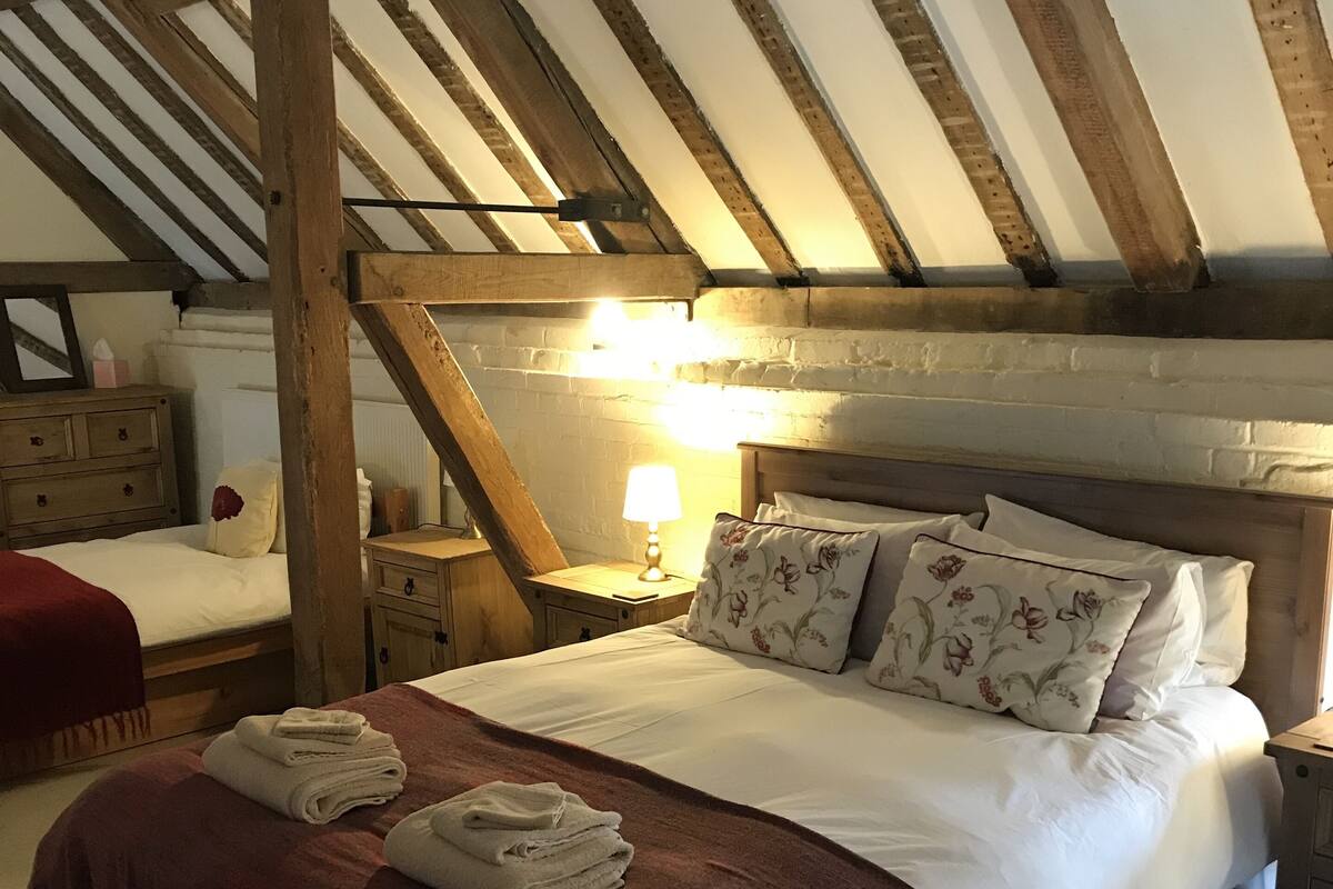 A bedroom with a wooden ceiling, a bed with white bedding and floral pillows, a nightstand with a lamp, and a red throw blanket.
