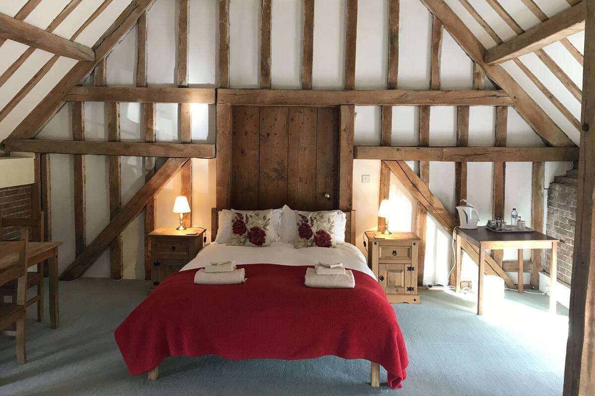 A bedroom with a sloped ceiling, wooden beams, a bed with red and white bedding, two bedside tables with lamps, and a small desk with a kettle.