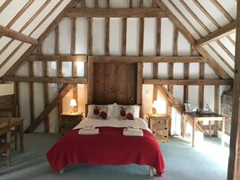 Une chambre avec un plafond en pente, des poutres en bois, un lit avec une literie rouge et blanche, deux tables de chevet avec des lampes et un petit bureau avec une bouilloire.