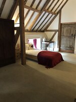 Un dormitorio con techo inclinado, vigas de madera, una cama con una colcha roja, una mesita con una jarra y una ventana con cortinas.