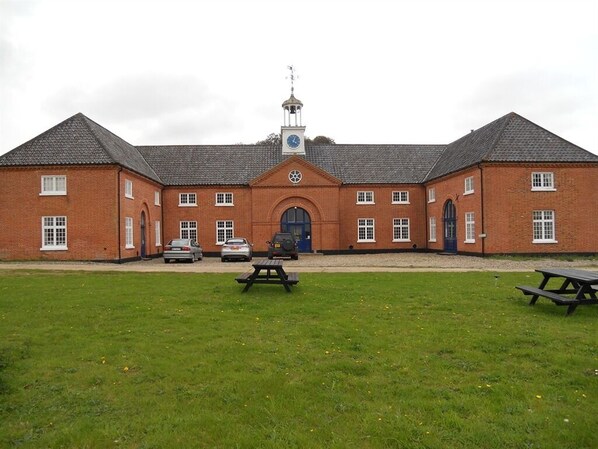 A large brick building with a central clock tower, multiple windows, and picnic tables in the foreground.
