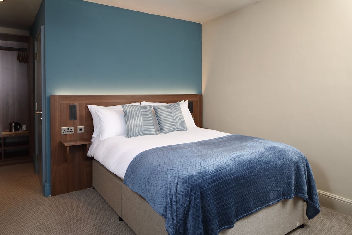 Un lit bien fait, avec une couverture bleue, une tête de lit en bois et une table de chevet surmontée d’une lampe.
