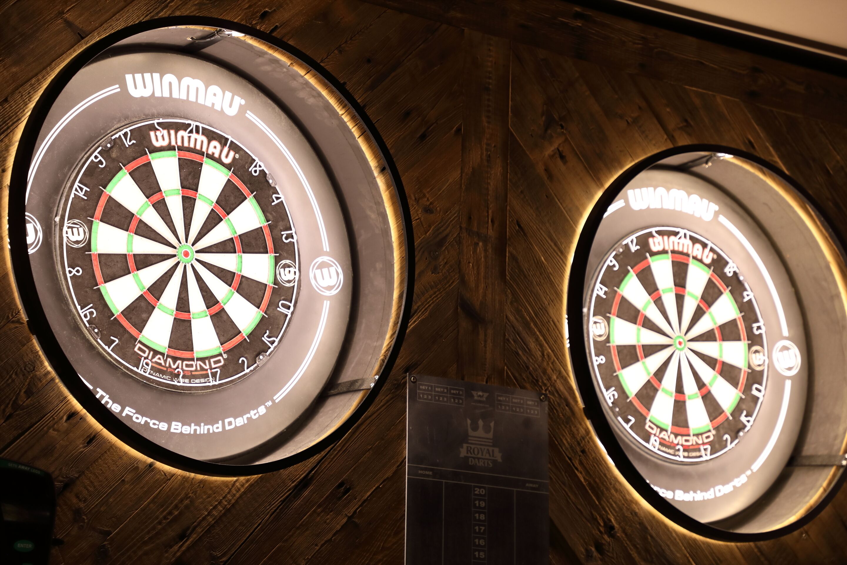Two illuminated dartboards mounted on a wooden wall.