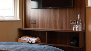 A hotel room with a bed, a TV mounted on the wall, a shelf with towels and glasses, and a window. - The Victoria Hotel (Burnham-on-Sea)