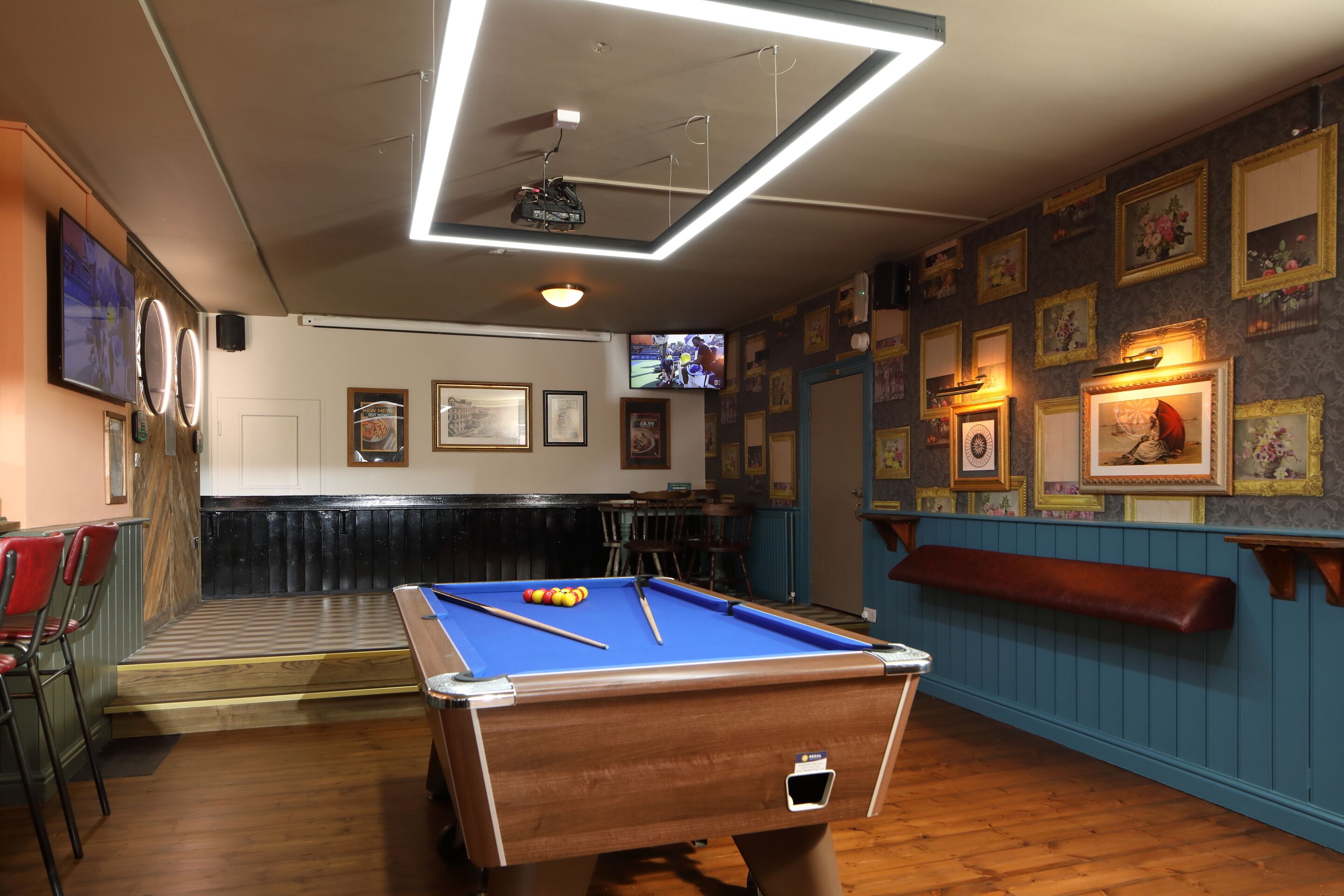 A pool table in a room with framed pictures on the walls, a television, and a bar area.