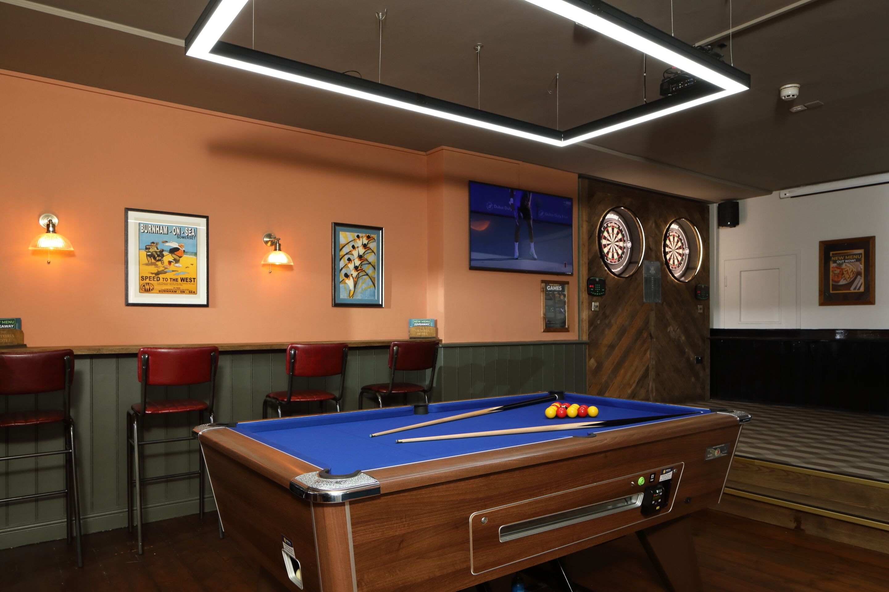 Une table de billard avec des billes, deux cibles de fléchettes et des affiches encadrées accrochées au mur.