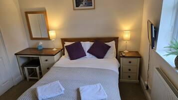 A neatly made bed with two towels, a bedside table with a lamp, a small desk, a mirror, and a framed picture on the wall.