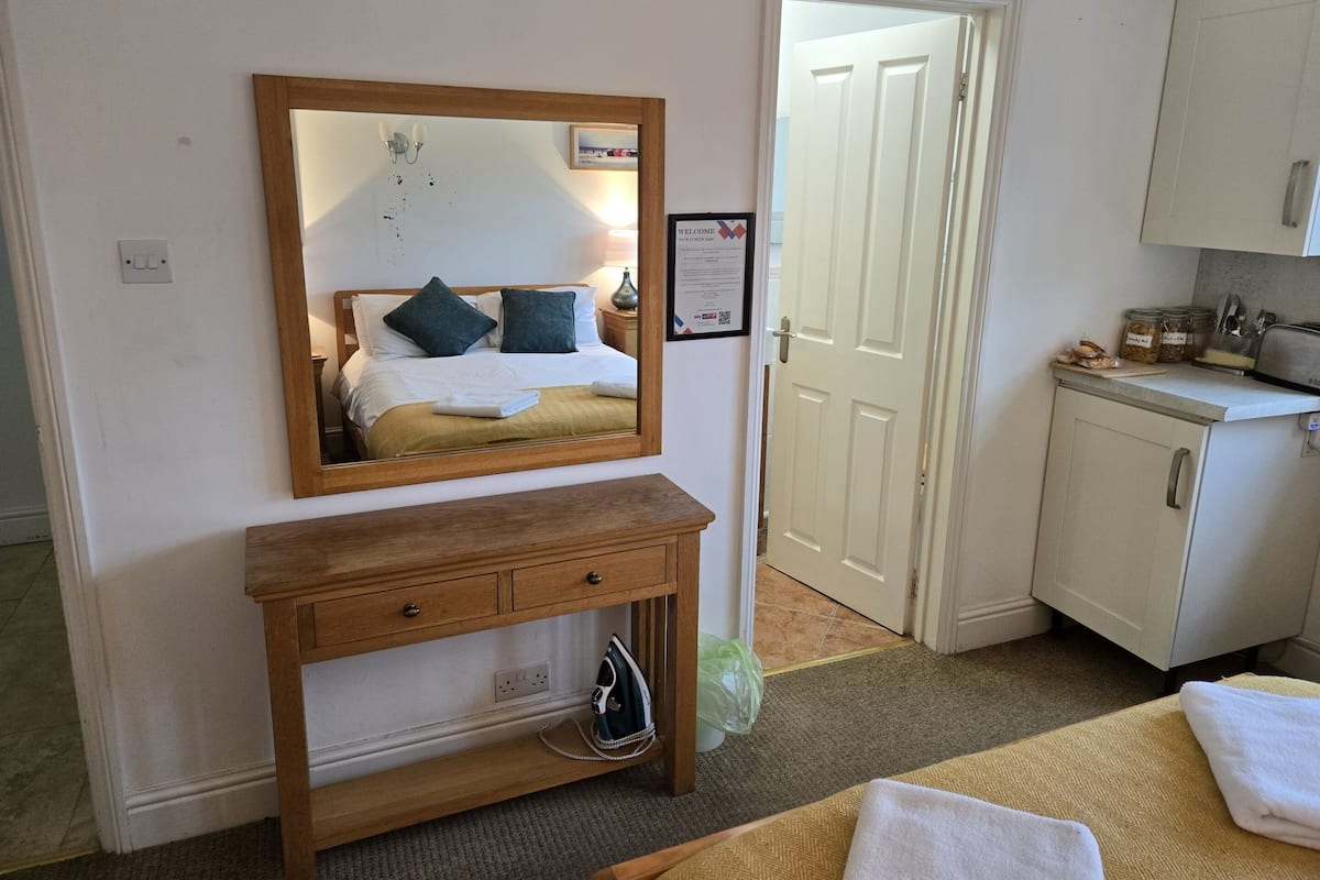 Une chambre d’hôtel comprenant un lit, une table de chevet en bois avec des tiroirs, un miroir, une porte donnant accès à la salle de bain et un coin cuisine équipé d’un grille-pain et de bocaux.