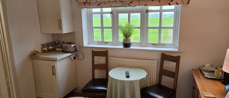 A compact hotel room with a kitchenette, a small dining table, a bed, and a window with a view of a green field.
