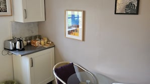 A compact kitchen with a toaster, kettle, and bread on the counter. There is a wicker chair with a purple cushion and a glass table. - Wayside (Norwich)