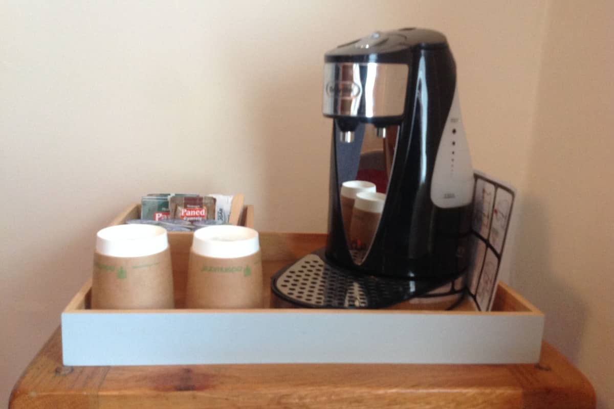 A coffee maker on a wooden tray with two paper cups and a box of Paned coffee pods.