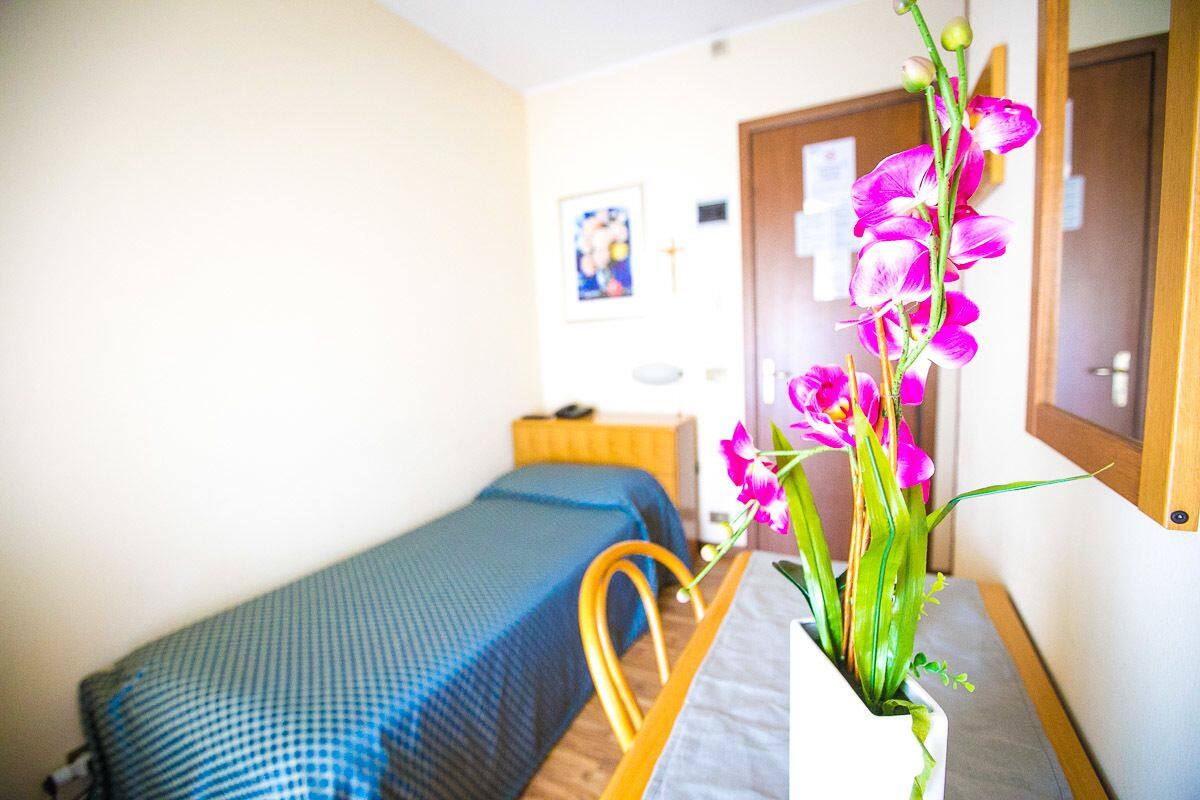 A small room with a single bed, a wooden chair, and a vase with flowers.