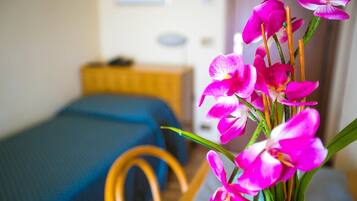 A bed with a blue cover, a wooden chair, and a vase with pink flowers in a room.