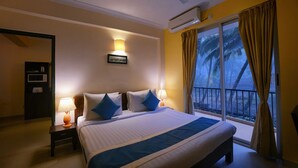 Habitación de hotel con una cama grande, dos mesitas de noche con lámparas, un equipo de aire acondicionado, vista a palmeras por la ventana y una puerta a la izquierda.