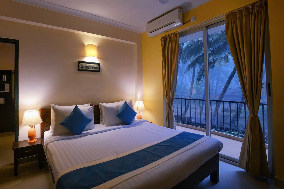 A hotel room with a large bed, two bedside tables with lamps, an air conditioning unit, a view of palm trees through the window, and a door to the left.