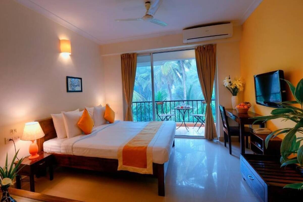 A hotel room with a bed, bedside table with a lamp, a TV, a balcony with a table and chairs, and a large window with curtains.