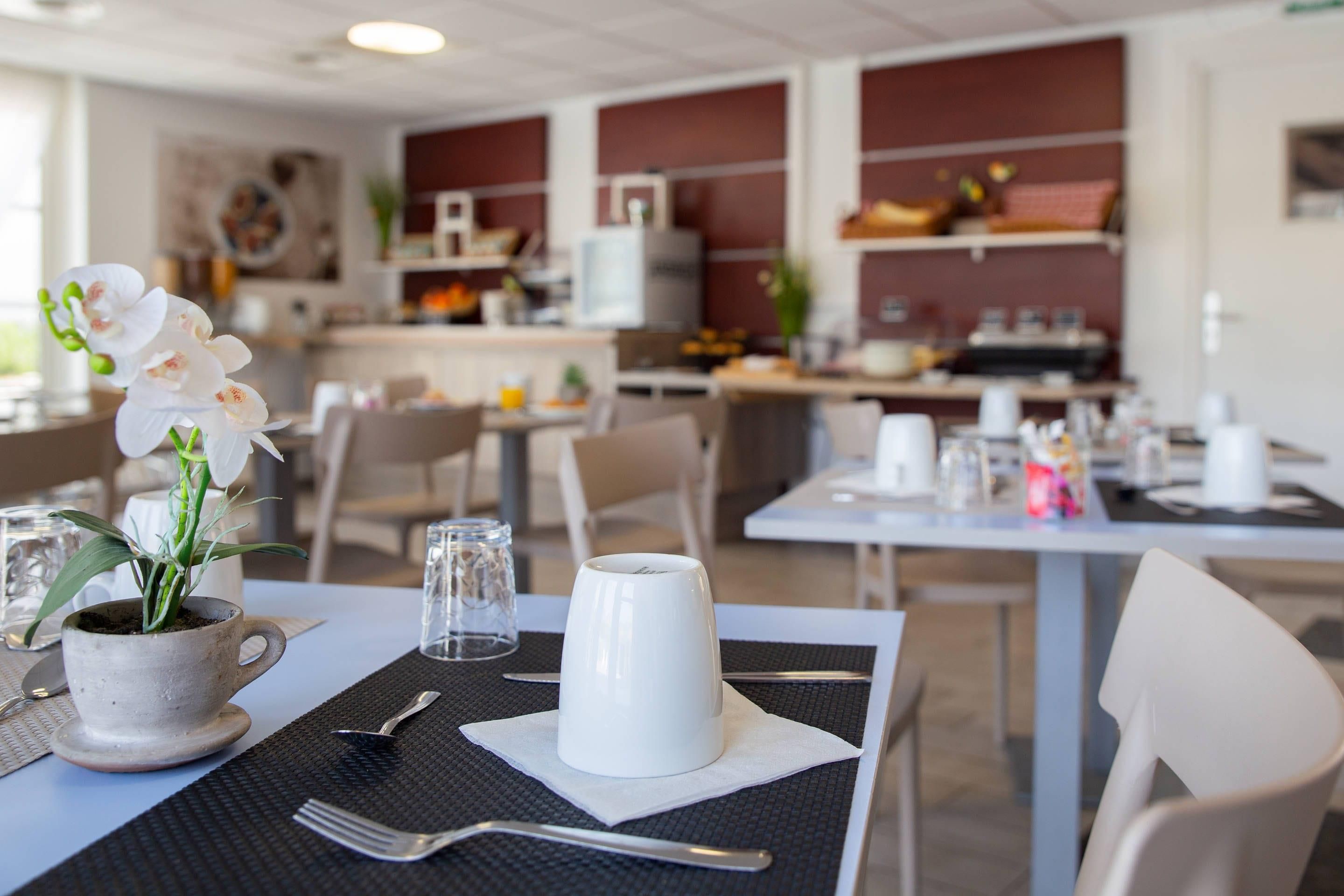 Un coin repas avec des tables préparées pour deux personnes, une orchidée blanche dans un pot en céramique et une tasse blanche posée sur une serviette.