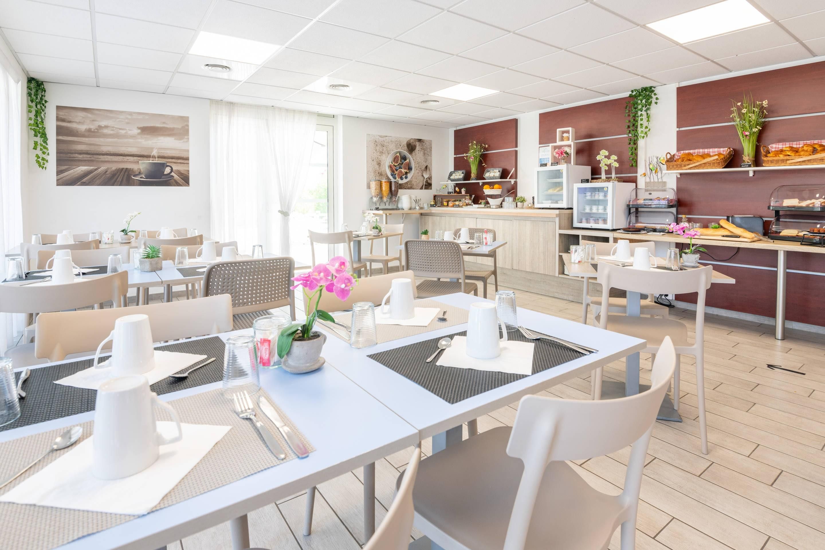 A modern hotel dining area with multiple tables, chairs, and a kitchen area in the background.