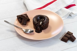 Une assiette contenant un fondant au chocolat, une cuillère et des morceaux de chocolat, le tout posé sur une surface en bois blanche.
