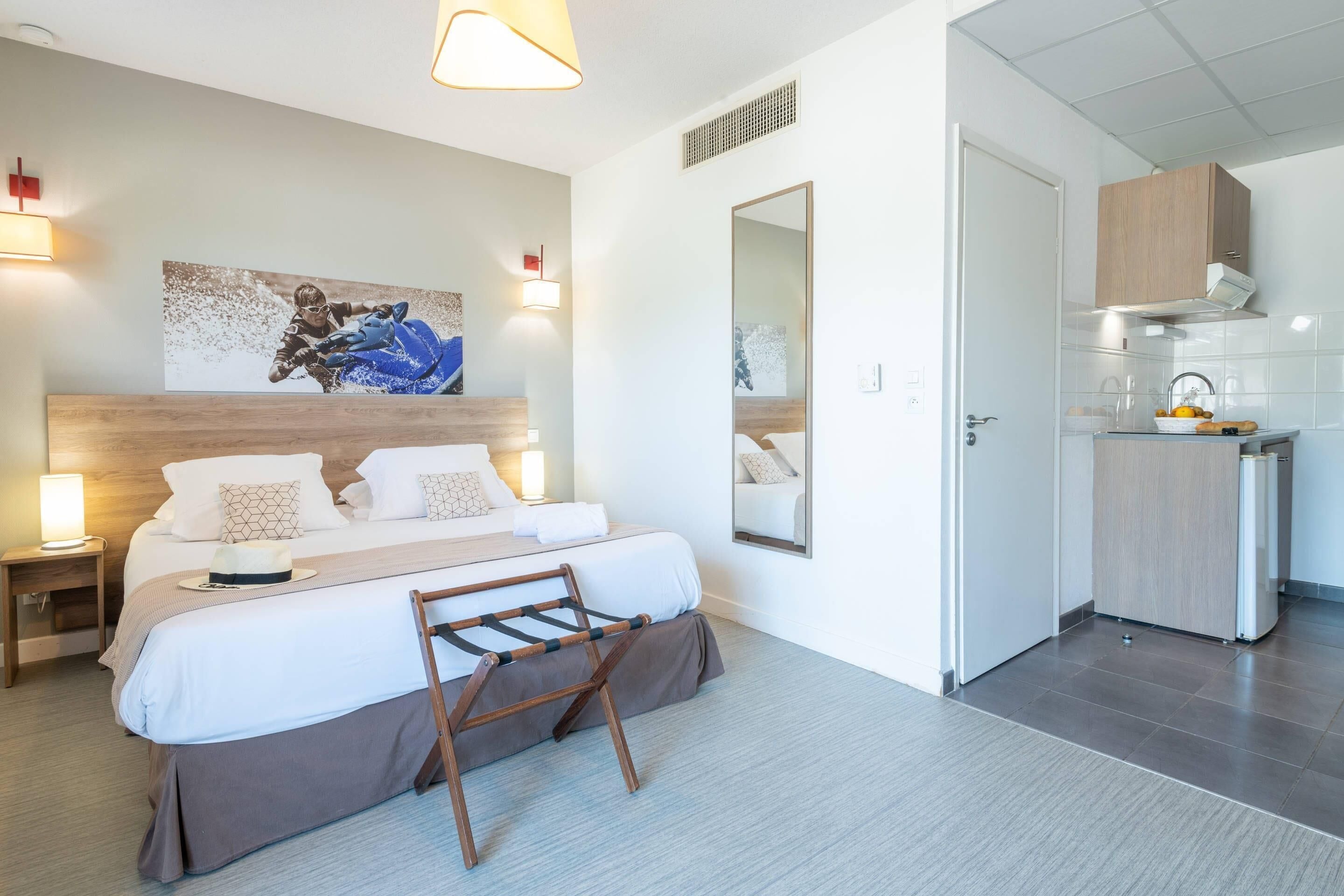 Una habitación de hotel moderna con una cama grande, lámparas de noche, un cabecero de madera, un espejo, una pequeña kitchenette y una obra de arte colgada en la pared.