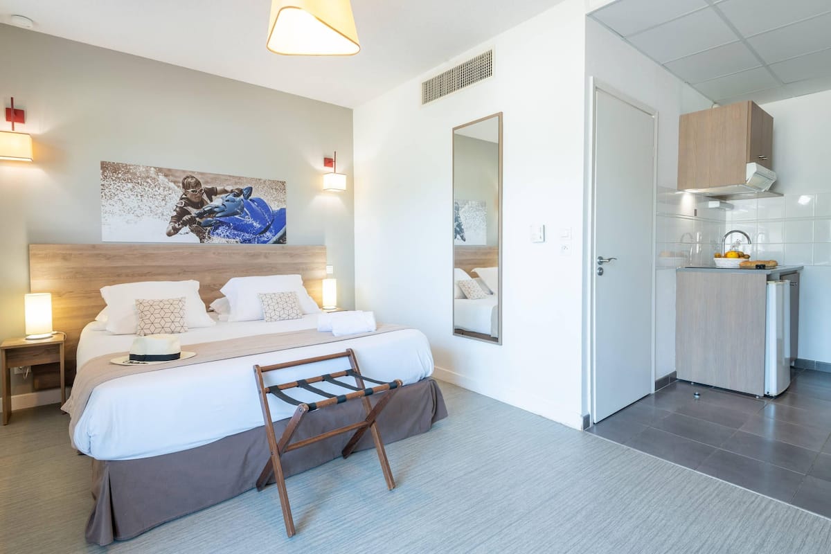 Una habitación de hotel moderna con una cama grande, lámparas de noche, un cabecero de madera, un espejo, una pequeña kitchenette y una obra de arte colgada en la pared.