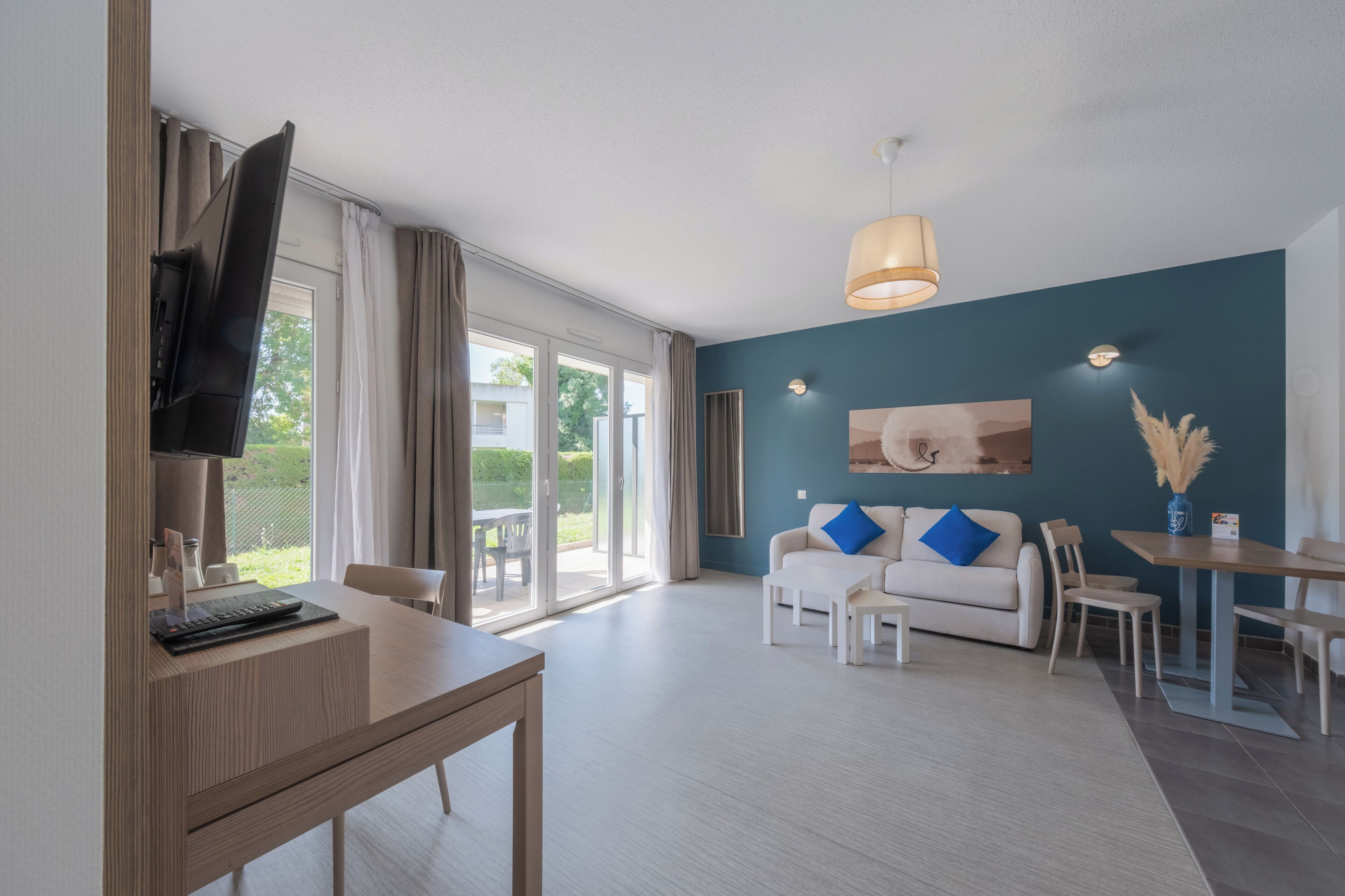 A modern living room with a flat-screen TV, a wooden dining table, a sofa with blue cushions, and a large window with curtains.
