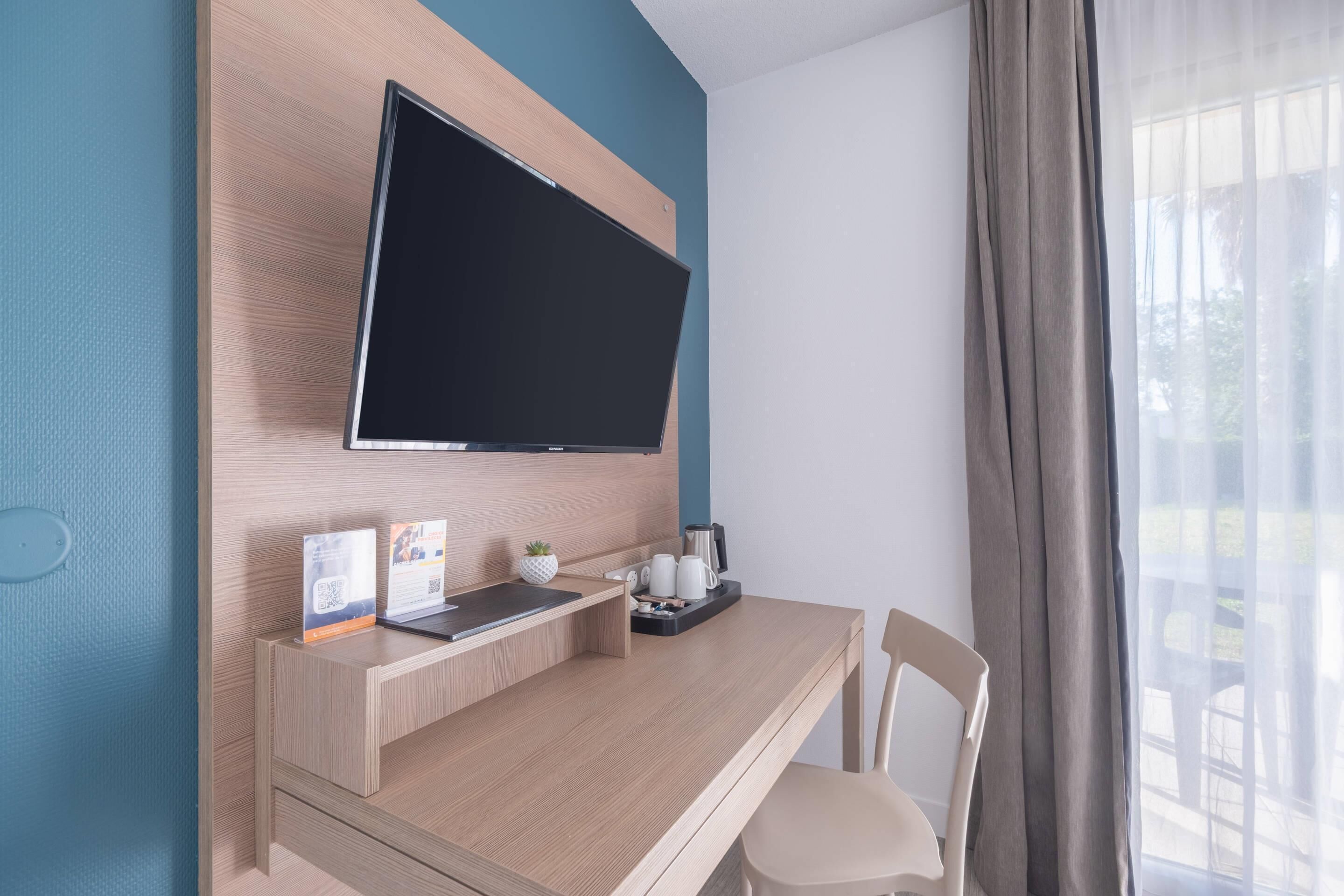 Una habitación moderna con una televisión de pantalla plana montada en una pared de madera, un escritorio con una computadora portátil, una cafetera y una planta pequeña.
