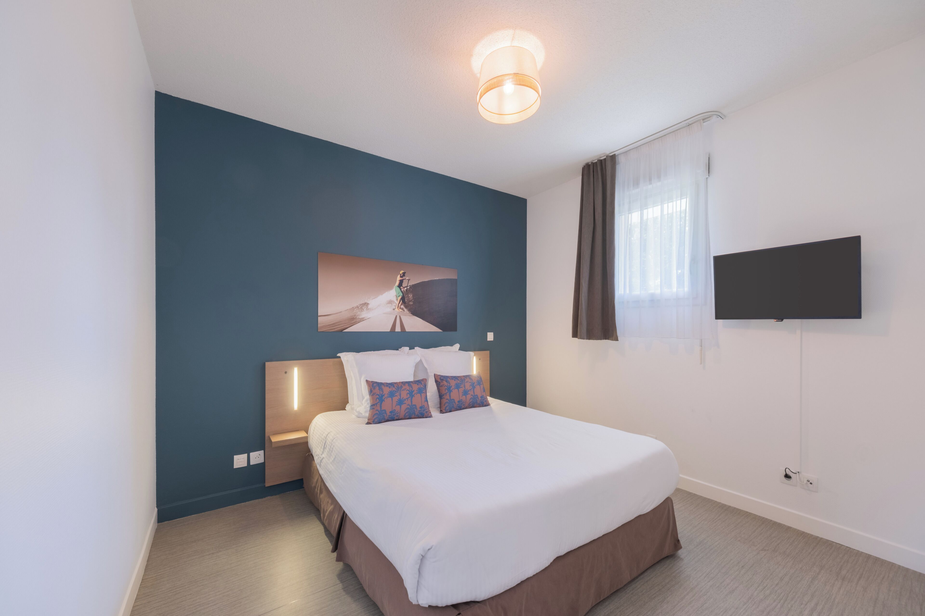 A hotel room with a bed, a mounted TV, a wall-mounted light fixture, and a painting.
