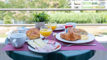 Une table de petit-déjeuner avec des croissants, du fromage, du jambon et du jus, installée sur un balcon offrant une vue sur les bâtiments et la verdure.