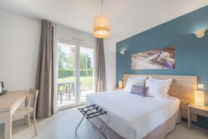 Ein Hotelzimmer mit Bett, Schreibtisch, Stuhl, Nachttisch mit Lampe und Blick ins Freie.