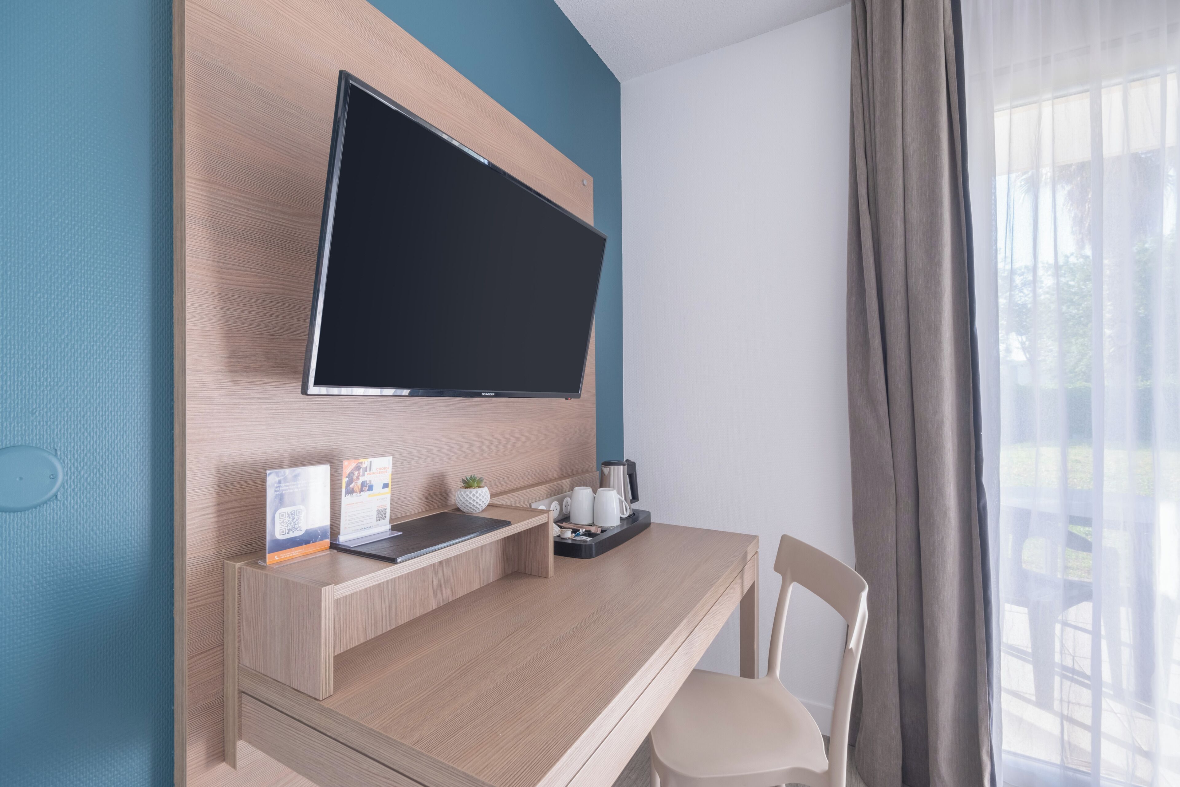 Habitación de hotel moderna con una pared de madera, un televisor de pantalla plana, un escritorio con una computadora portátil, una cafetera y una planta pequeña.