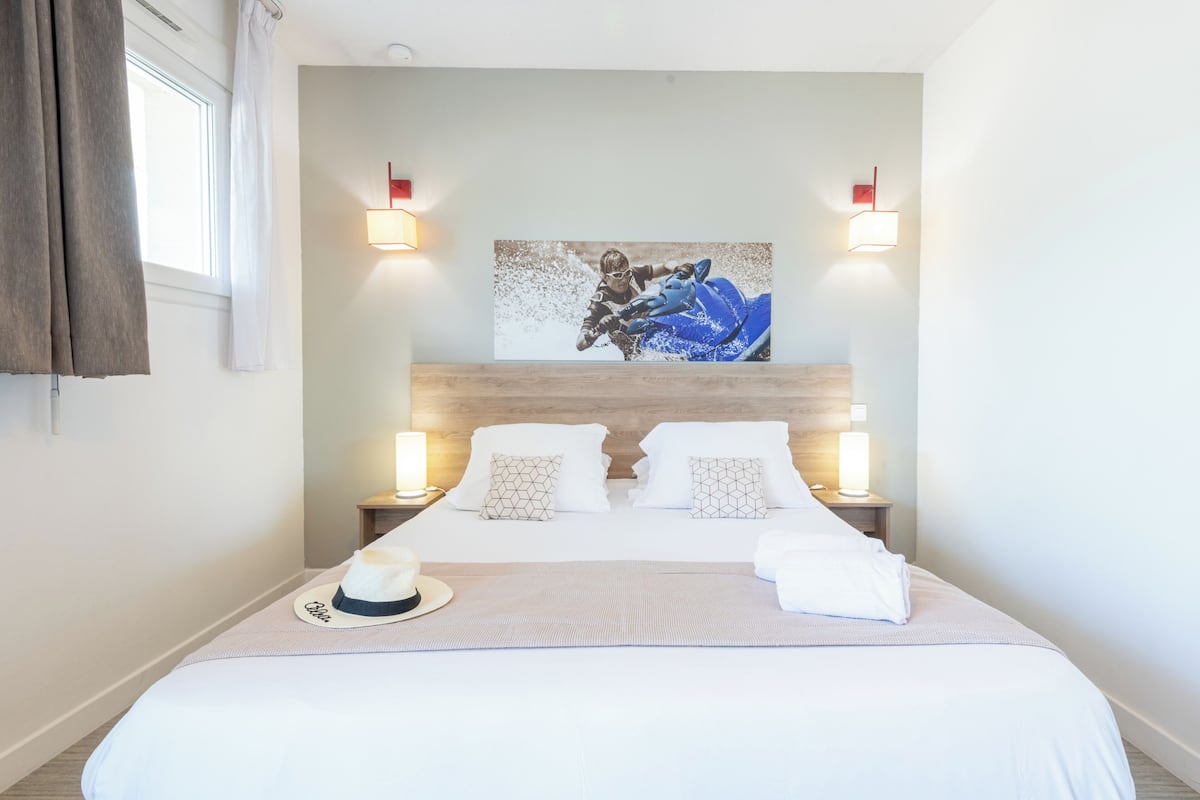 Una cama bien hecha con cubrecama blanco y almohadas, una obra de arte en la pared y dos lámparas de noche.