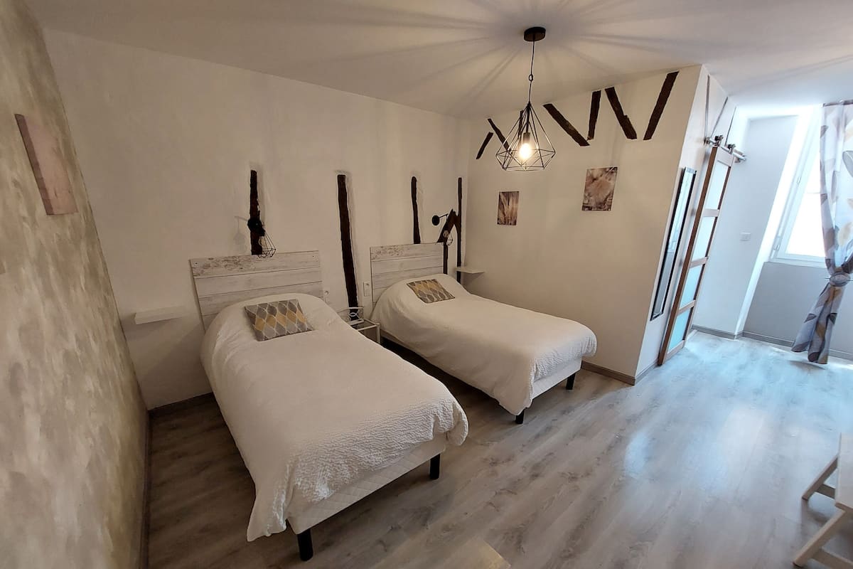 A bedroom with two single beds, wooden flooring, a hanging light fixture, and a window with curtains.