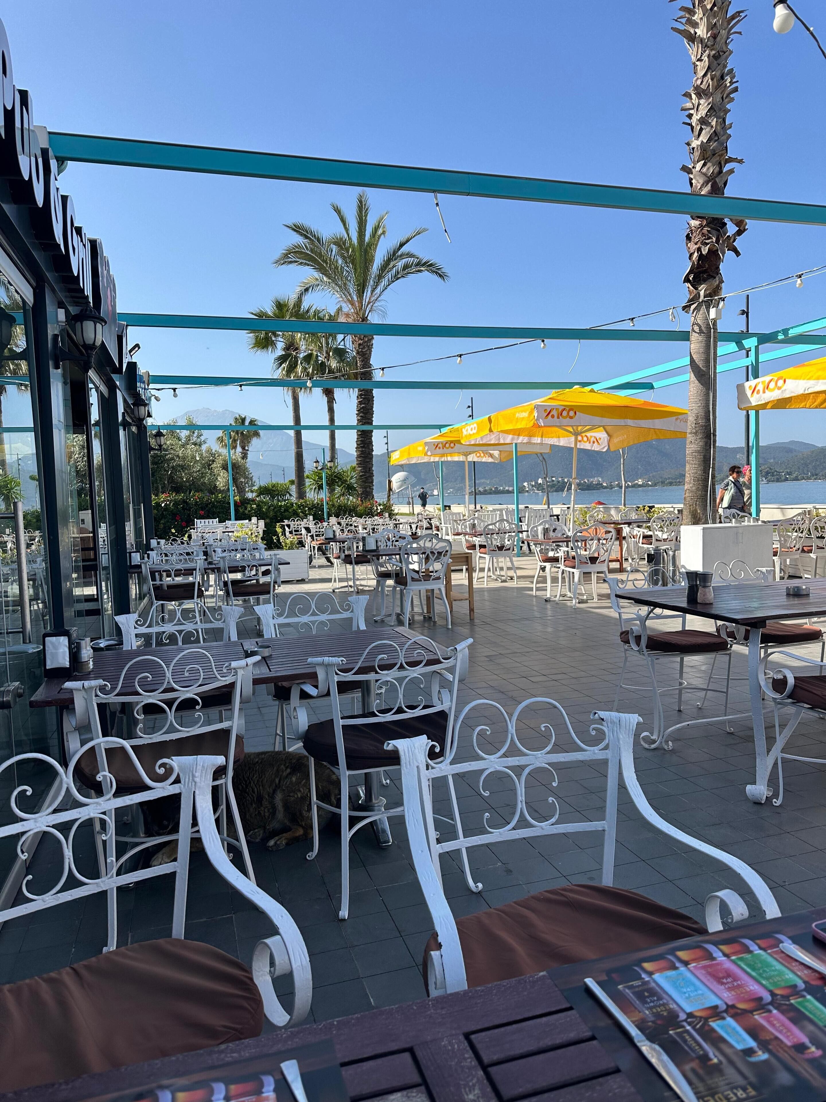 An outdoor cafe with white metal chairs and tables, yellow umbrellas, and a view of the sea and mountains in the background.
