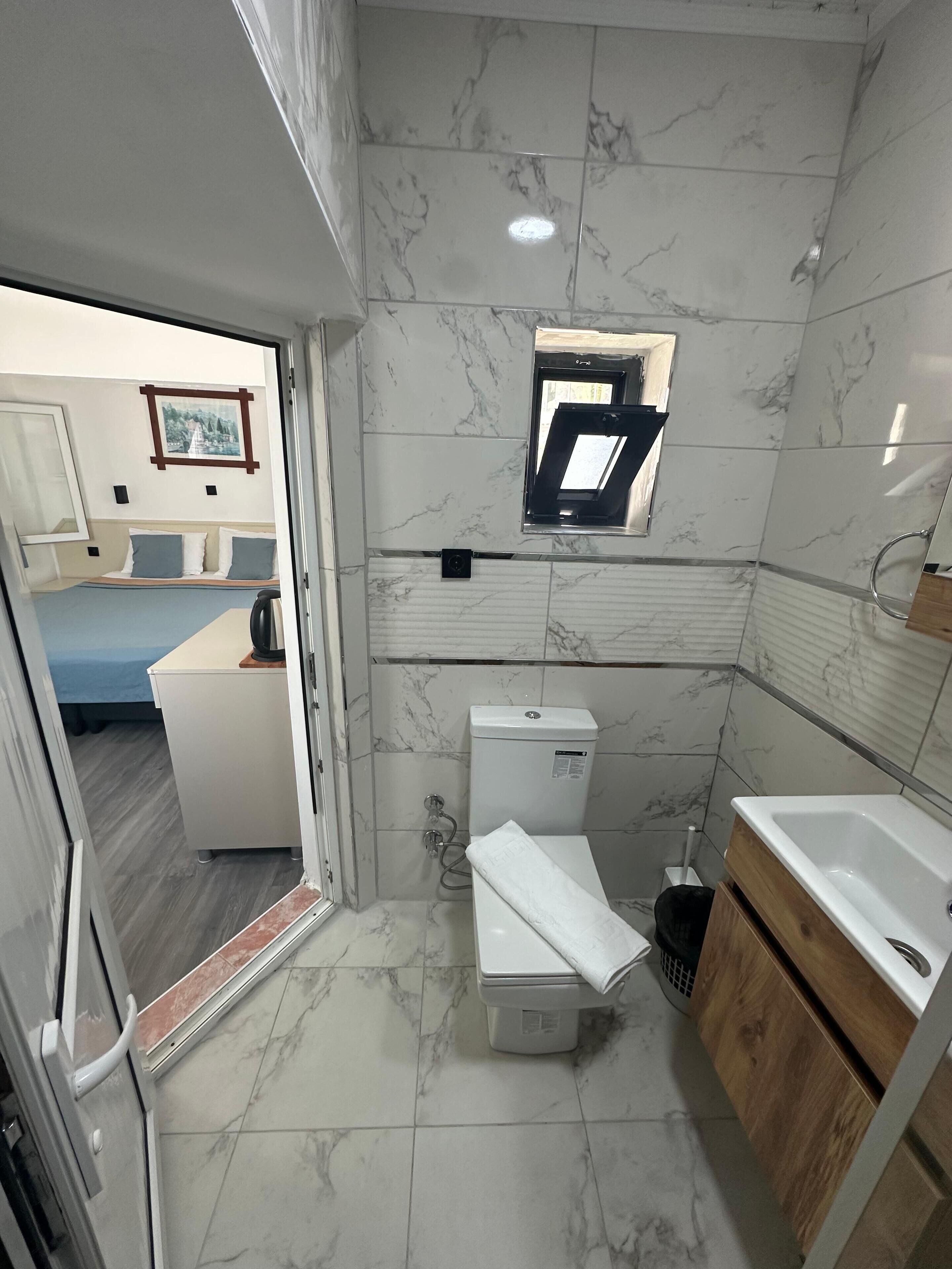 A compact bathroom with a toilet, sink, and shower area, featuring marble tiles and a small window.