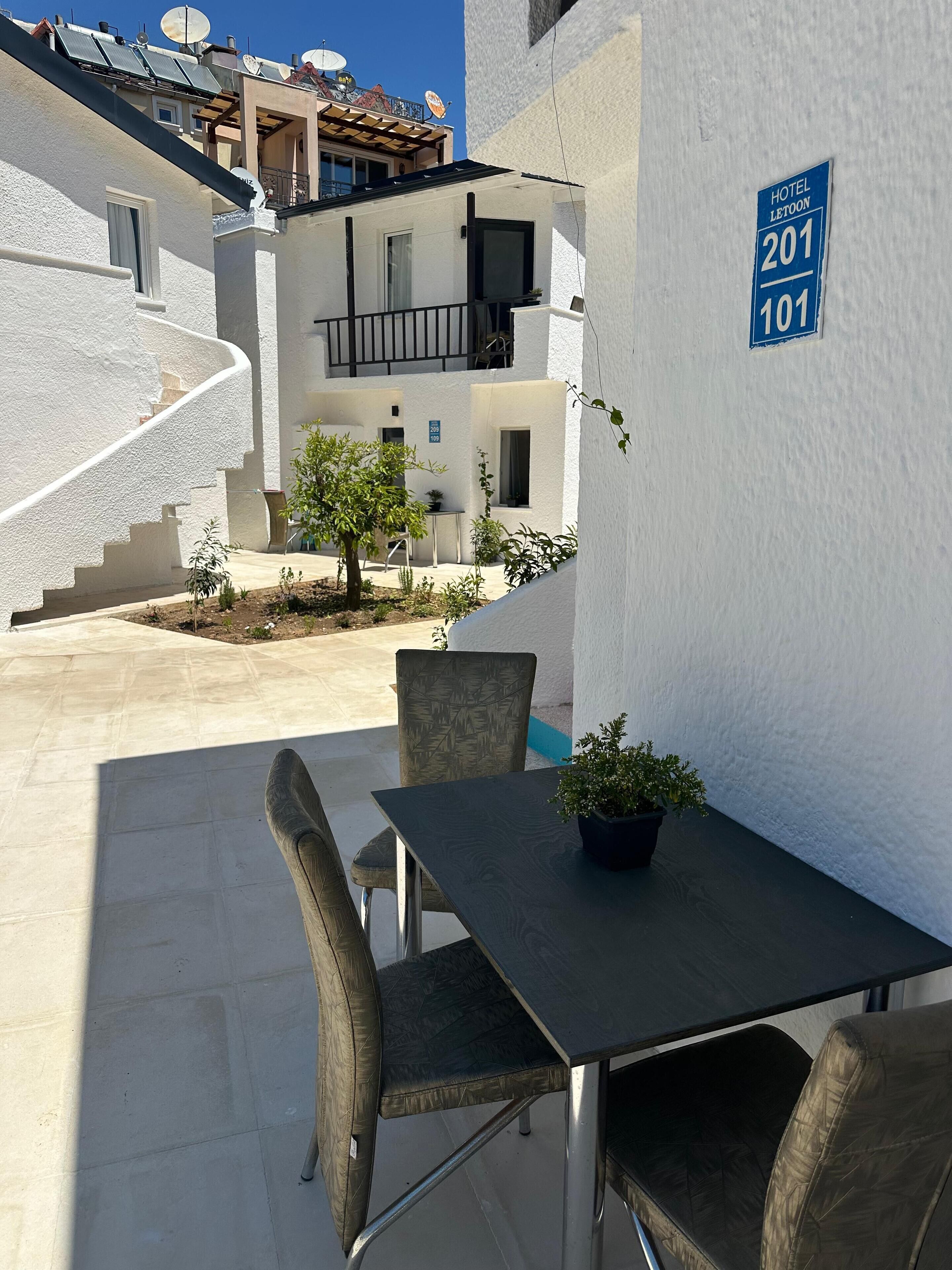 Une cour avec une table et des chaises, un pot de fleurs et un bâtiment sur lequel est apposée une enseigne indiquant « Hôtel Letoon », avec les numéros de chambre 201 et 101.